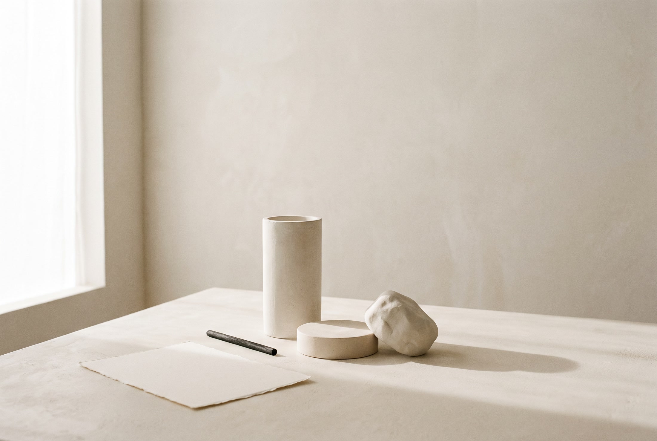 A ceramic vessel, a disc of raw clay, and a torn sheet of paper arranged on a sunlit studio table.